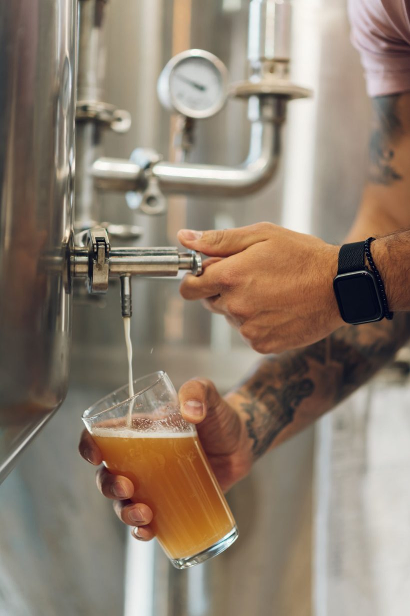 Close shot of a man worker examining beer glass while working at the brewery. Brewer working in his craft brewery.Focus on a man hands filling up glass of beer.